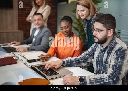 A diverse group of people is seated around a table, engrossed in their work on laptops. They appear focused and busy, possibly collaborating on projec Stock Photo