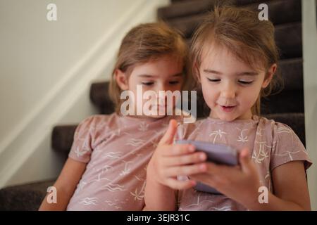 Two preschool age girls using a smartphone sitting on stairs at home Stock Photo