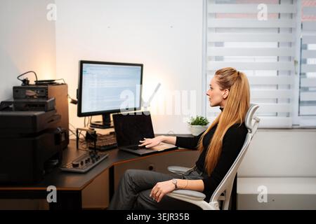 Young adult woman concentrating while using computer and laptop at home desk Stock Photo