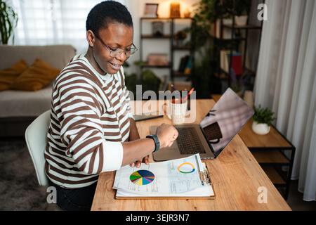 Young adult woman analyzing data on clipboard and checking smartwatch at home office Stock Photo