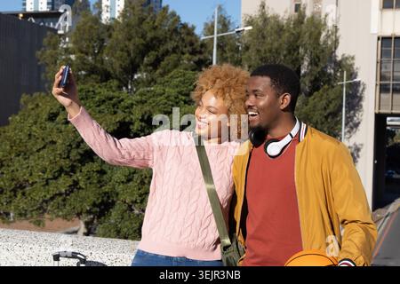 African American couple taking smartphone selfie at overlook with suitcase, man holding basketball. Duo, lifestyle, outdoor, urb Stock Photo