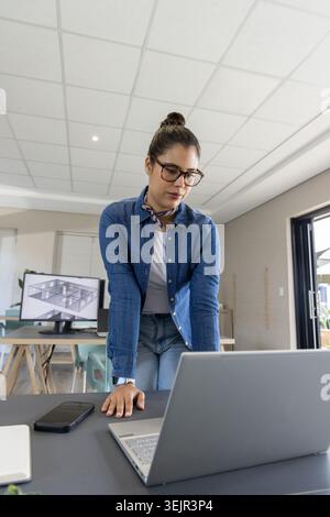 Woman in denim shirt leaning over laptop in office checking phone and reviewing floorplan on screen. Design, architecture, plann Stock Photo