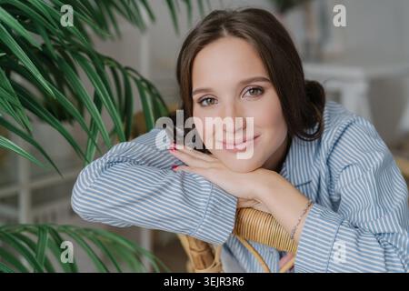 Close up shot of pretty dark haired young woman has charming gentle smile, leans on hands, poses indoor near green plantation, h Stock Photo