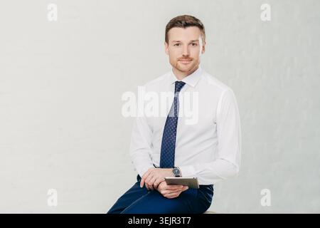 Isolated shot of successful male manager wears elegant white shirt, black trousers and tie, uses modern tablet computer for work Stock Photo