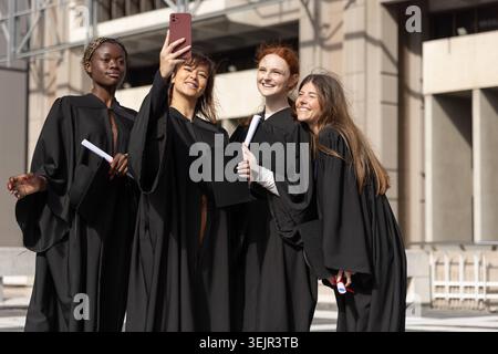 Diverse female friends taking selfie with pink smartphone in black gowns holding diplomas on plaza. Outdoor, campus, mortarboard Stock Photo