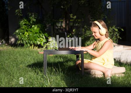 Child girl in headphones   is using a laptop and communicates on the Internet  outdoor. homeschooling, distant learning Stock Photo