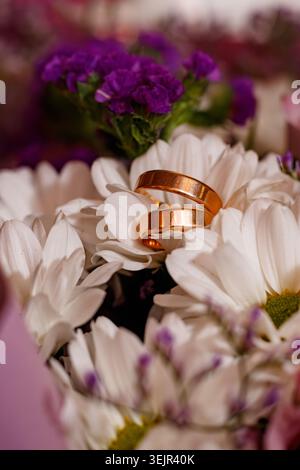 Two shiny gold rings sit on top of white daisy petals surrounded by small purple flowers in a bouquet. The arrangement shows a mix of bright colors an Stock Photo