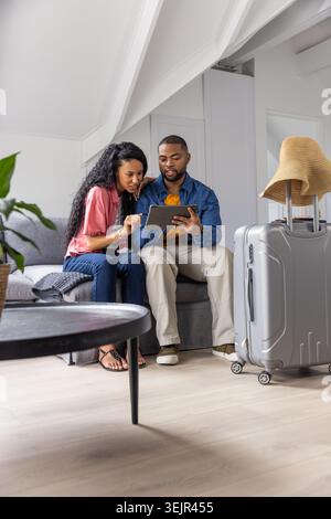 Diverse couple sitting on gray sofa in loft, studying tablet planning trip near suitcase with hat. Luggage, duo, travel, itinera Stock Photo
