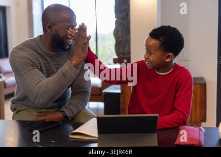 African American father and son high-fiving at home counter near window, using tablet and notebook. Family, together, parenting, Stock Photo