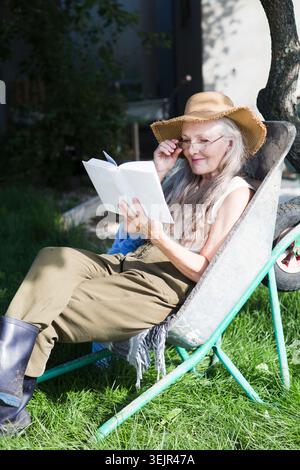 Elderly woman  with long grey hair  sits in a garden wheelbarrow and and reads a book in the summer garden near the house. Stock Photo