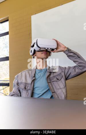 Asian man sitting at classroom table adjusting white VR headset with headphones near window. Tech, immersive, desk, denim, teal, Stock Photo