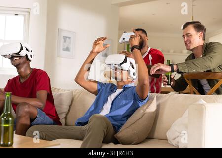 Diverse men watching player wearing VR playing game after controller lift with beer in living room. Headset, sofa, beverage, lifestyle, casual, modern Stock Photo