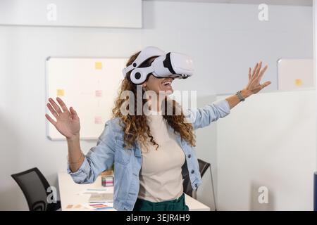 Woman wearing white VR headset and denim jacket, reaching out, smiling at desk with color swatches. Immersive, futuristic, techn Stock Photo