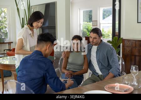 Diverse couple consulting two advisors, reviewing paperwork on wooden dining table with pen. Home, daylight, interior, greenery, Stock Photo