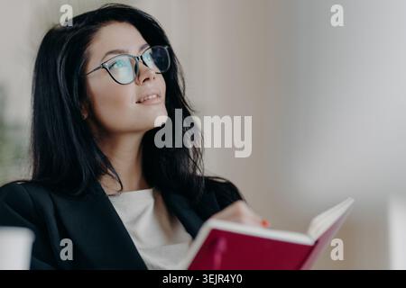 Indoor shot of thoughtful dreamy brunette female jouranlist works on book review, holds opened notepad, writes down some ideas o Stock Photo