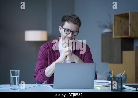 Pensive young German man home office employee sits at desk at workplace operates online on laptop Stock Photo