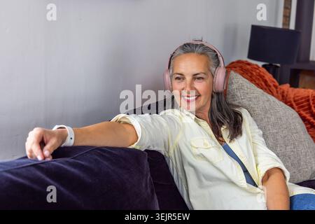Senior woman reclining on dark purple sofa in living room, wearing pink headphones, reaching. Retiree, cozy, home, purple, comfort, relaxation, music Stock Photo