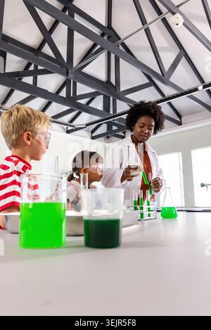 African American teacher in lab coat guiding children with goggles pipetting green liquid at lab. Instructor, pupils, classroom, Stock Photo