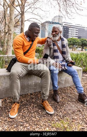African American male friends, senior sitting on bench at park holding phones in orange sweatshirt. Smartphones, plaza, urban, overcast, casual, outdo Stock Photo