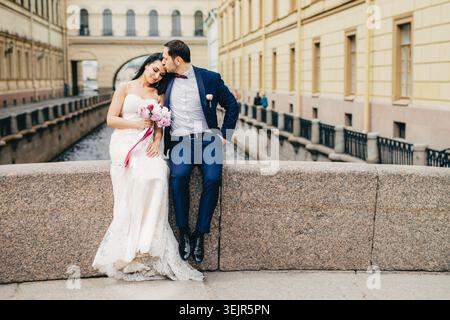 Affectionate married couple sit on bridge togetherm enjoy fresh air, have good relationship, kiss each other. Young female in wh Stock Photo