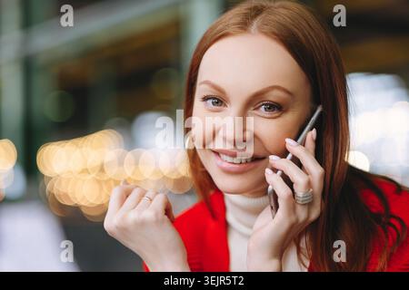 Close up shot of attractive carefree female student talks on mobile phone, enjoys conversation with groupmate, has pleasant smile, poses over blurred Stock Photo