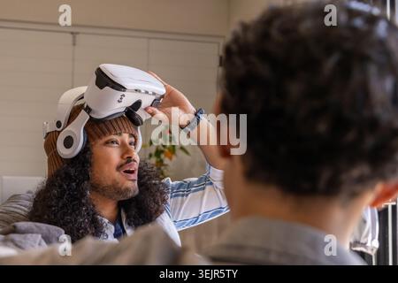 Male friends sitting on sofa, one lifting white VR headset with headphones, wearing beanie. Virtual, reality, gadget, home, livi Stock Photo