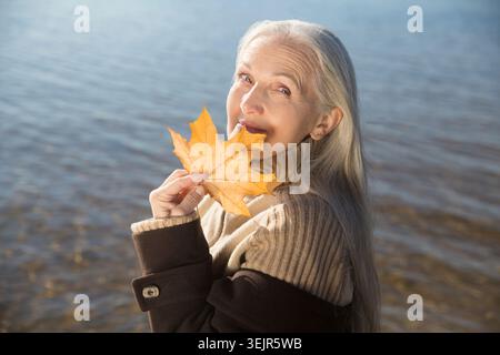 Elderly woman with gray hair wearing a brown coat holds a yellow maple leaf in his hand on the bank of the river. Slow life. Enj Stock Photo