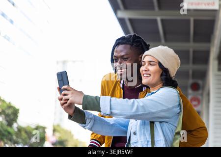 Female and African American male friends leaning at transit shelter taking selfie with smartphone. Urban, duo, candid, daylight, Stock Photo