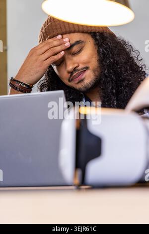 Man rubbing forehead, sitting at desk in home office wearing beanie with laptop and VR headset. Workspace, computer, goggles, la Stock Photo