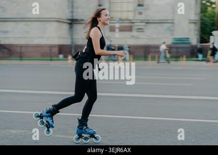 Rest leisure and lifestyle concept. Cheerful dark haired young European woman breathes fresh air while rollerskating has hobby l Stock Photo