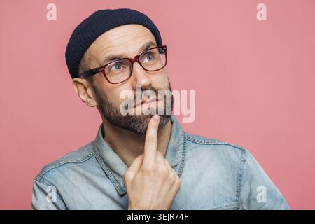 Thoughtful bearded man in glasses looking up and touching chin on pink background Stock Photo