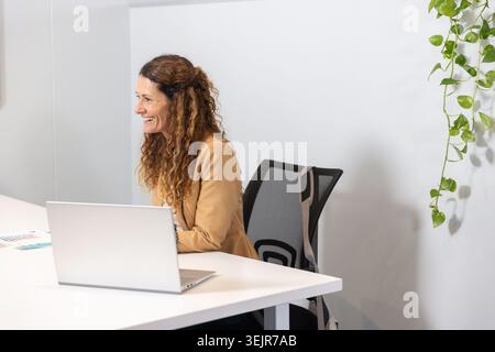 Mature woman wearing tan blazer sitting in office using silver laptop with brochure and plant. Workspace, professional, minimalist, ergonomic, greener Stock Photo