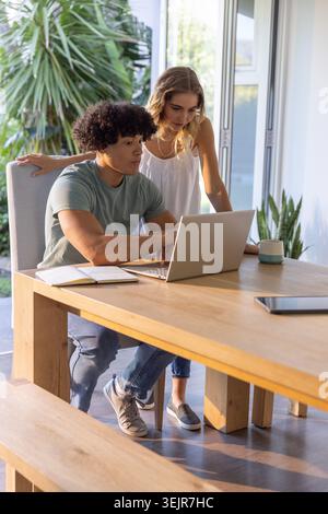 Diverse couple working at wooden dining table using silver laptop while being surrounded by plants. Homeoffice, collaboration, d Stock Photo