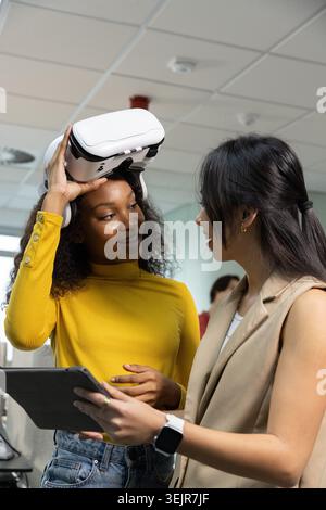 Diverse female coworkers adjusting white VR headset and holding tablet in modern office. Collaboration, training, technology, de Stock Photo