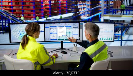 Two Workers Analyze Ecommerce Orders On Multiple Screens In Busy Warehouse. Stock Photo