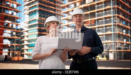 Two Construction Inspectors Review Building Plans On Laptop At Site. Stock Photo