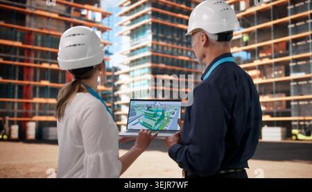 Two Architects Review BIM Model On Laptop While Planning Construction Site. Stock Photo