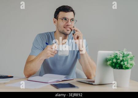 Man talking on smartphone while working from home with laptop and coffee Stock Photo