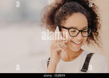 Smiling young woman talking on smartphone wearing glasses indoors Stock Photo