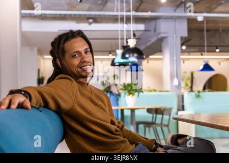 African American man in late twenties sitting on blue bench in coworking lounge checking wristwatch. Timepiece, workspace, casua Stock Photo