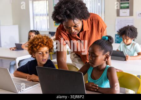 African American teacher wearing rust-orange shirt leaning over students using laptop at classroom. Collaborative, mentoring, te Stock Photo