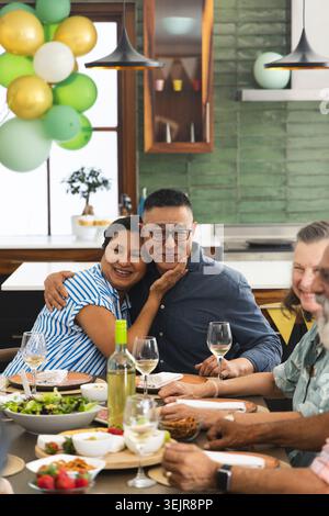 Diverse friends sharing celebratory meal at home on wooden dining table with wine and balloons. Cozy, intimate, gathering, family, kitchen, warmth, ru Stock Photo