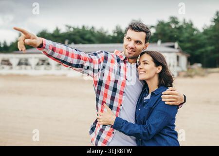 Couple hugging on beach with man pointing toward new home near seaside Stock Photo