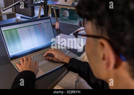 Asian man typing spreadsheet on laptop in shared office desk area with pink headphones, copy space. Coworking, metrics, graphs, Stock Photo