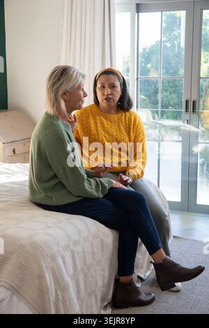 Mature female friends sitting on bed edge in softly lit bedroom, one gesturing, both wearing boots. Cozy, conversation, friendsh Stock Photo