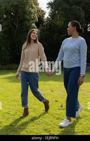 Diverse female partners walking hand in hand on sunlit grassy lawn, wearing sweaters and watches. Couple, outdoors, park, garden, greenery, nature, ro Stock Photo