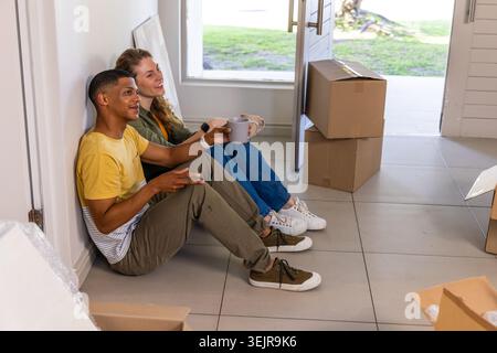 Diverse couple sitting on tiled floor near open front door, holding mugs and unpacking boxes. Partners, moving, relocation, entryway, sunlit, minimali Stock Photo