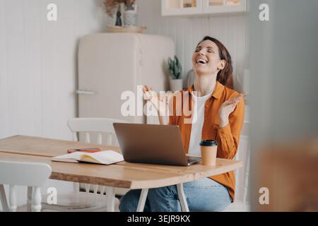 Woman celebrating success while using laptop at kitchen table working from home Stock Photo