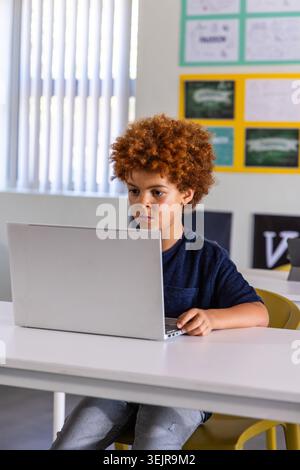 African American child boy sitting on yellow chair at white desk in classroom using silver laptop. School, computer, focused, learning, daylight, post Stock Photo