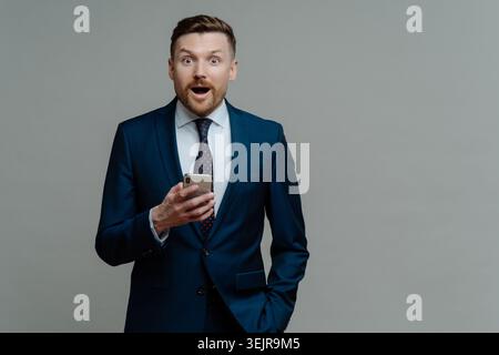 Business man checking smartphone with shocked expression on gray studio background Stock Photo